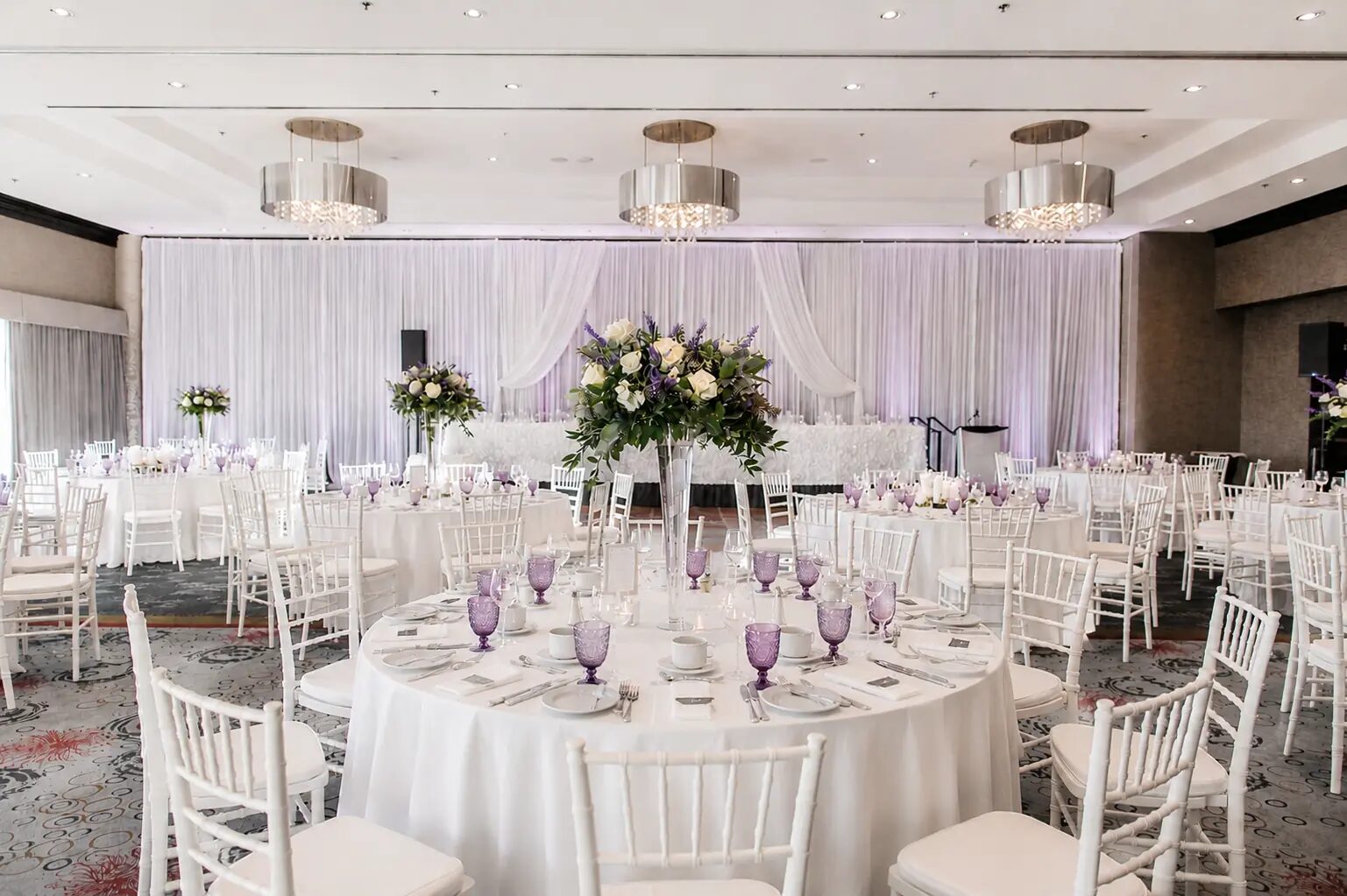 Wedding reception in the ballroom with white draping, chiavari chairs and tall floral centrepieces